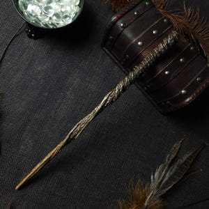 May include: A long, slender wand featuring a dark, twisted handle that transitions into a lighter, wood-grained shaft. The handle has a textured, spiraled design with metallic highlights, while the tapered tip remains smooth and light brown.