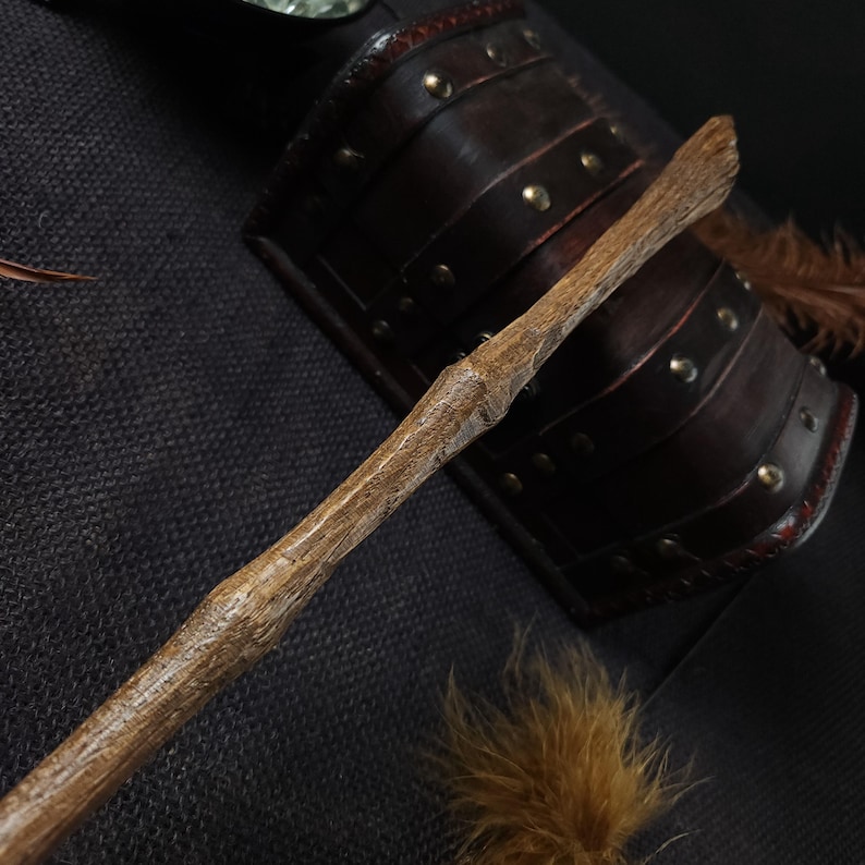 May include: A handcrafted wooden wand with a textured surface, featuring a natural, light brown color. The wand is positioned diagonally, with a dark brown leather bracer in the background, adorned with brass studs. A cluster of brown feathers adds detail.