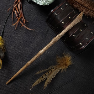 May include: A handcrafted wooden wand with a natural, textured finish. The wand is light brown and tapers to a point. Decorative feathers and leather wrist cuffs are also visible, suggesting a fantasy or cosplay theme.