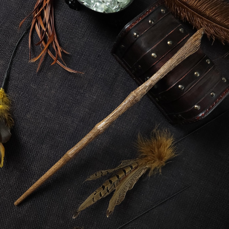 May include: A handcrafted wooden wand with a natural, textured finish. The wand is light brown and tapers to a point. Decorative feathers and leather wrist cuffs are also visible, suggesting a fantasy or cosplay theme.