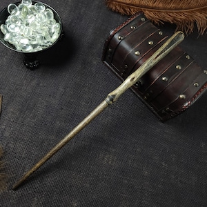 May include: A wooden wand with a dark brown finish and a light green handle. The wand is lying on a dark surface next to a small black bowl filled with clear glass beads and a brown leather chest.
