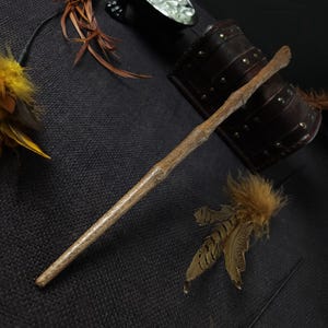 May include: A handcrafted wooden wand with a natural, light brown finish. The wand is tapered and has a textured, branch-like appearance. Decorative feathers and a leather cuff are also visible.