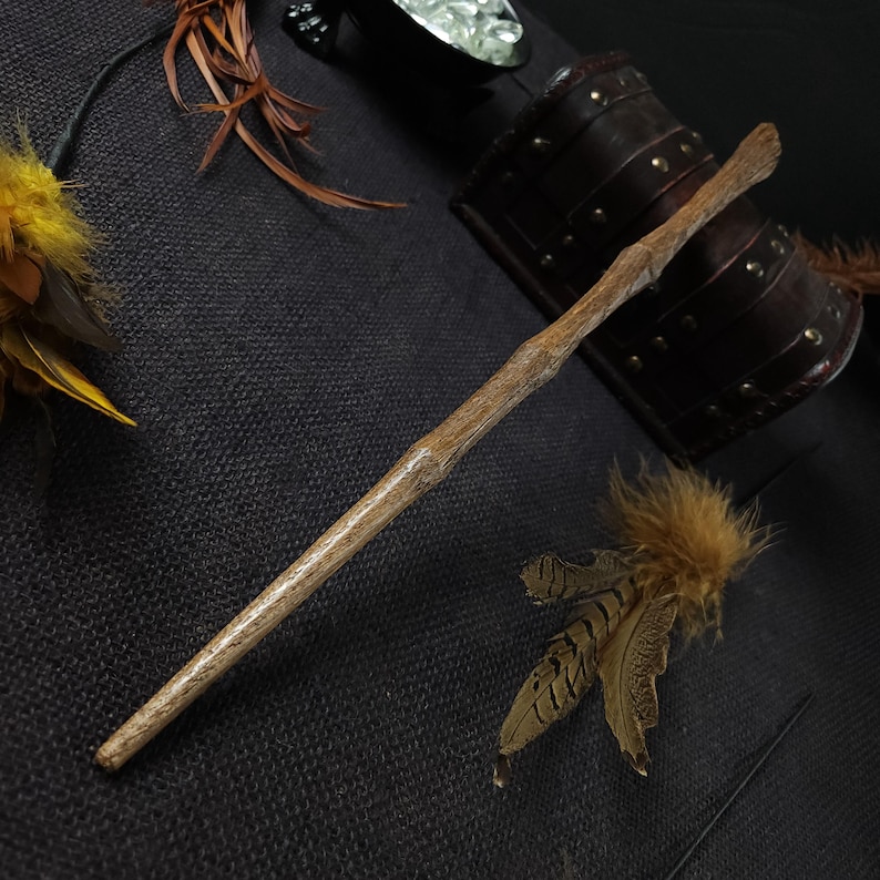 May include: A handcrafted wooden wand with a natural, light brown finish. The wand is tapered and has a textured, branch-like appearance. Decorative feathers and a leather cuff are also visible.