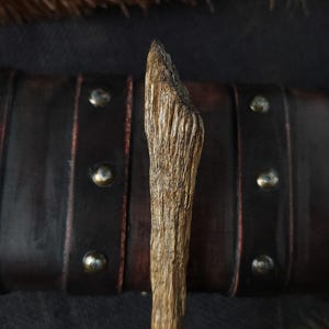 May include: A handcrafted wooden wand with a textured surface, featuring a natural, light brown color. The wand is set against a dark background with a leather strap and metallic studs, and a feather.