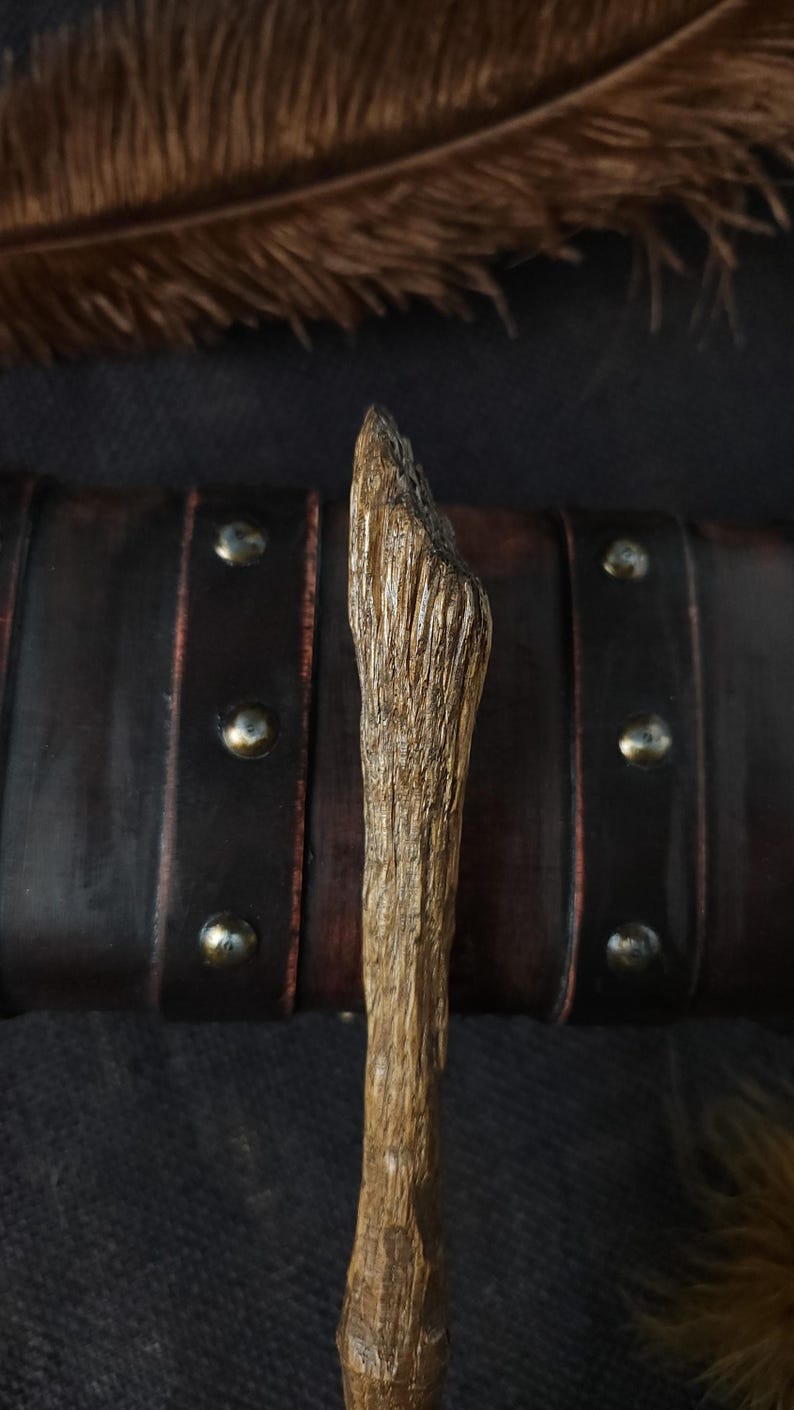 May include: A handcrafted wooden wand with a textured surface, featuring a natural, light brown color. The wand is set against a dark background with a leather strap and metallic studs, and a feather.
