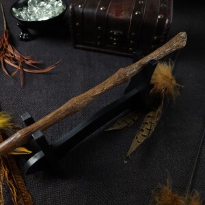 May include: A handcrafted wooden wand with a textured, natural finish, resting on a black stand. The wand is brown with a detailed, organic design. Decorative feathers and a small treasure chest are in the background, suggesting a fantasy or wizarding theme.