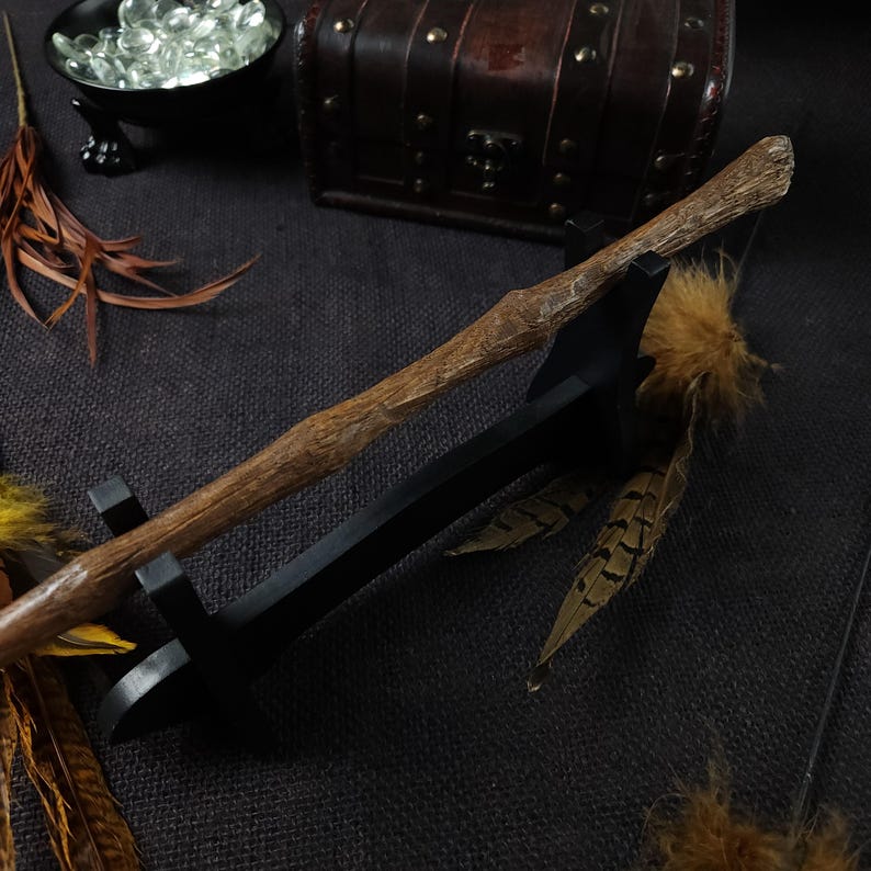 May include: A handcrafted wooden wand with a textured, natural finish, resting on a black stand. The wand is brown with a detailed, organic design. Decorative feathers and a small treasure chest are in the background, suggesting a fantasy or wizarding theme.