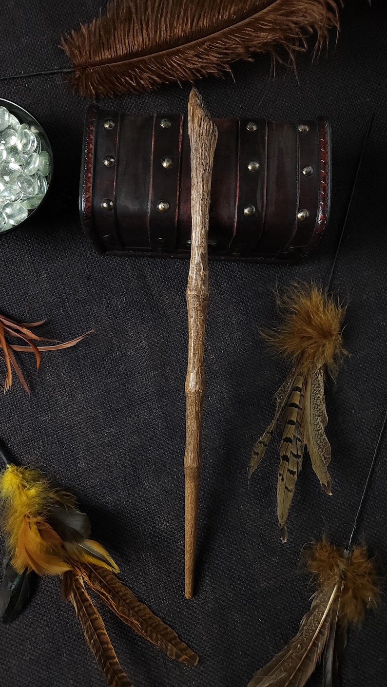 May include: A wooden wand with a textured surface, positioned vertically. The wand is surrounded by decorative feathers, a small leather chest with metal studs, and a bowl of clear glass beads. The items are arranged on a dark, textured background.