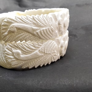 Puede incluir: Un brazalete blanco en forma de bangle con intrincados tallados de pavos reales y plumas.