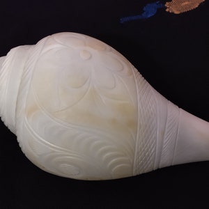 Op de afbeelding: Een grote, gebroken witte schelp met ingewikkelde bloemen en geometrische gravures. De schelp heeft een glad, gepolijst oppervlak en een natuurlijke, organische vorm. De gravures voegen een decoratief element toe.