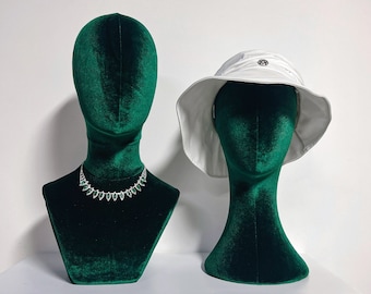 May include: Two green velvet mannequins, one wearing a necklace with emerald green stones and the other wearing a white sun hat.