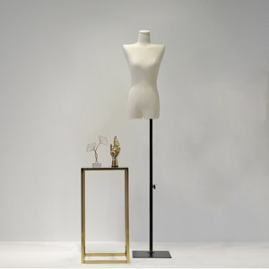 May include: A cream-coloured dress form on a black adjustable stand. A small gold-coloured hand statue and a wire sculpture of leaves sit on a gold-framed table. The background is a plain white wall.