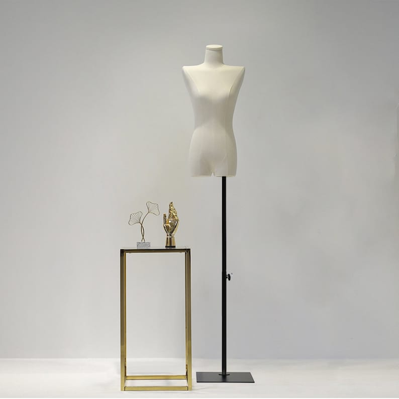 May include: A cream-coloured dress form on a black adjustable stand. A small gold-coloured hand statue and a wire sculpture of leaves sit on a gold-framed table. The background is a plain white wall.