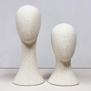 Puede incluir: Dos cabezas de maniquí cubiertas de tela, una alta y otra baja, de pie una al lado de la otra. Las cabezas son de forma ovalada con un cuello largo y una base acampanada. La tela tiene un patrón moteado gris claro y blanco, creando una apariencia texturizada. El fondo es gris claro liso.