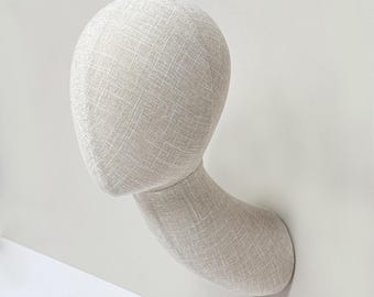 May include: A neutral-toned mannequin head and neck, designed for display. The head is oval-shaped, and the neck curves gracefully. The surface has a textured, woven appearance. The mannequin is mounted on a white wall, suggesting a retail or studio setting.