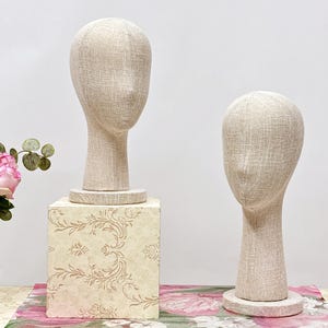 Op de afbeelding: Twee beige stoffen hoofdmankins. Eén mannequin staat op een decoratieve vierkante standaard met een bloemenpatroon. De andere mannequin staat op een ronde basis. De achtergrond is wit met een roze bloemenpatroon.