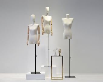 May include: Three white dress forms with adjustable black stands. Two forms have articulated wooden arms. A small gold and black display table holds decorative objects. The background is a plain white.