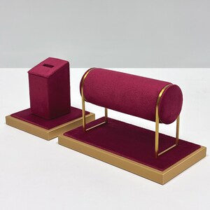 May include: Two jewelry display stands. One is a burgundy velvet angled box. The other is a burgundy velvet cylinder on a gold metal frame. Both are on gold rectangular bases. The stands are for displaying bracelets, necklaces, and rings.