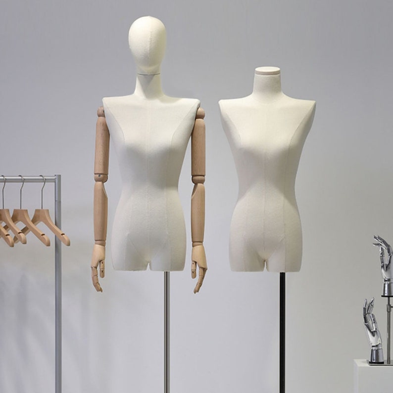 May include: Two cream-coloured dressmaker mannequins on black stands. One has articulated wooden arms and a head, the other is a torso form. A clothing rack with wooden hangers is in the background.