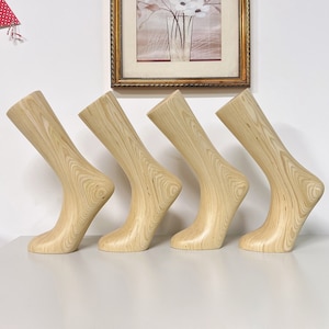 May include: Four wooden sock mannequins, each in a different pose, are arranged in a row on a white surface. The mannequins are made of light-coloured wood with a natural grain pattern.