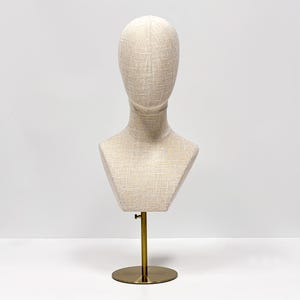 May include: A beige fabric mannequin head and bust form on a gold-colored metal stand. The bust has a textured surface and a round base. The stand is adjustable.