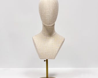 May include: A beige fabric mannequin head and bust form on a gold-colored metal stand. The bust has a textured surface and a round base. The stand is adjustable.