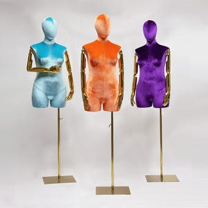May include: Three female mannequins with gold-colored arms and stands. The mannequins are made of velvet and are in different colors: light blue, orange, and purple.