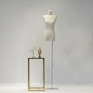 May include: A cream-coloured dress form on a silver stand, with a small gold and glass side table. The table has a gold frame and a black top, with two decorative gold sculptures on top.
