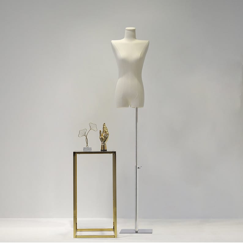 May include: A cream-coloured dress form on a silver stand, with a small gold and glass side table. The table has a gold frame and a black top, with two decorative gold sculptures on top.