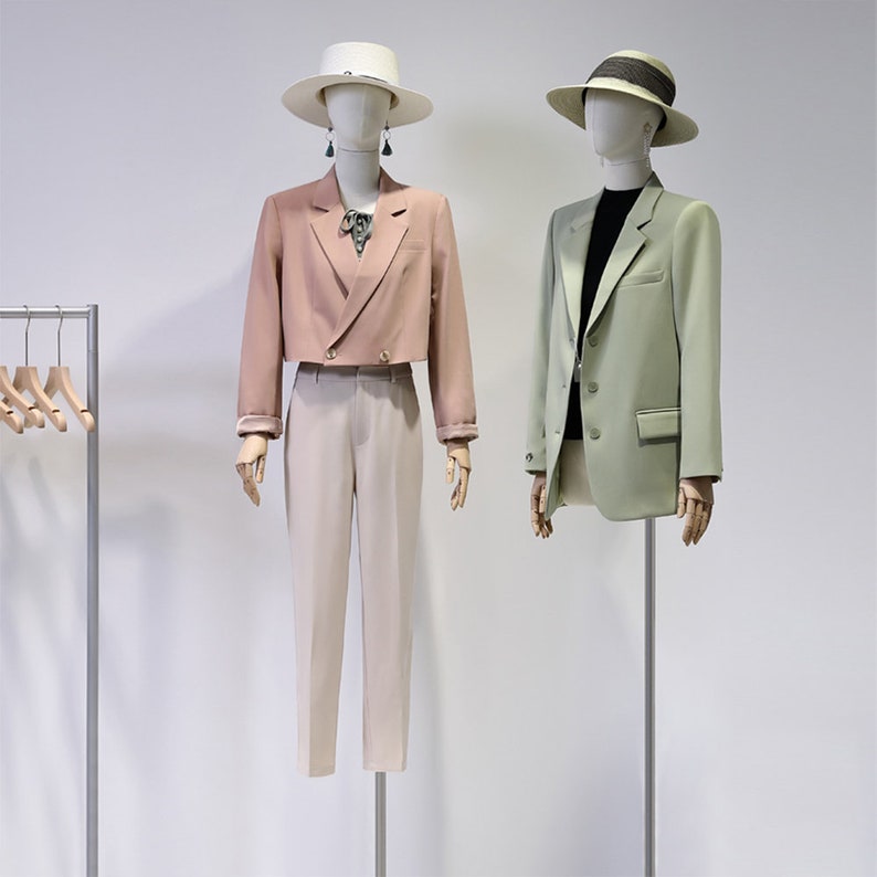 May include: Two mannequins showcase fashionable ensembles. One features a cropped blush blazer, beige trousers, and a white hat. The other is in a sage green blazer, black top, and a hat with a black band. Both outfits are accessorized with hats and earrings.