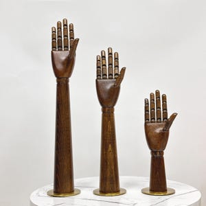 Könnte beinhalten: Drei hölzerne Hand-Mannequins mit ausgestreckten Händen, jeweils auf einem Messingsockel. Die Mannequins sind unterschiedlich groß, das größte links und das kleinste rechts.