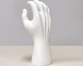 May include: A white hand sculpture with a curved index finger and open palm. The sculpture has a smooth, glossy finish and is set against a neutral background. The hand is mounted on a cylindrical base.