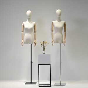 May include: Two dressmaker mannequins with articulated wooden arms and a cream-colored torso stand on adjustable metal poles. A small white display table with a black metal frame holds a vase of dried flowers. The background is a plain white wall.