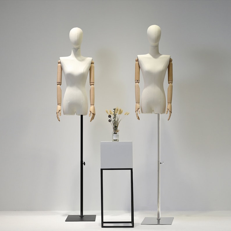 May include: Two dressmaker mannequins with articulated wooden arms and a cream-colored torso stand on adjustable metal poles. A small white display table with a black metal frame holds a vase of dried flowers. The background is a plain white wall.