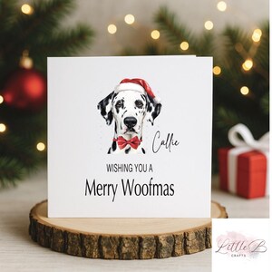 Może przedstawiać: Biała kartka świąteczna z dalmatyńczykiem w czapce Świętego Mikołaja i czerwoną muszką. Na kartce napis "Wishing You a Merry Woofmas" z imieniem "Callie" powyżej. Kartka jest prezentowana na drewnianym plastrze.