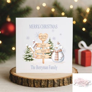Może przedstawiać: Kwadratowa kartka świąteczna z akwarelową ilustracją. Kartka przedstawia bałwana, drogowskaz z imionami oraz tekst "Merry Christmas" i "The Berryman Family". Drogowskaz głosi "2025" i ma imiona Daniel, Stacey i Olivia.
