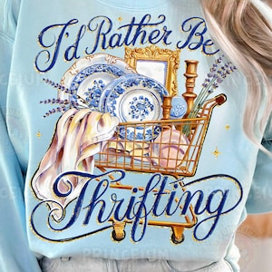 Op de afbeelding: Lichtblauw sweatshirt met de tekst "I'd Rather Be Thrifting" in een decoratief lettertype. De afbeelding bevat een winkelwagentje gevuld met vintage items zoals borden, een spiegel en lavendel. Het ontwerp heeft een vintage esthetiek.