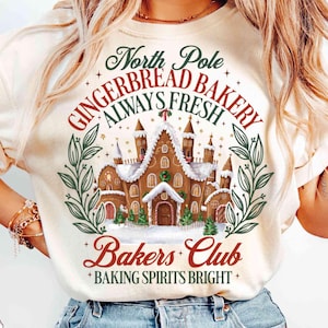 May include: Cream-colored t-shirt featuring a gingerbread house design with the text "North Pole Gingerbread Bakery Always Fresh Bakers Club Baking Spirits Bright." The design includes green and red accents, and a wreath around the bakery.