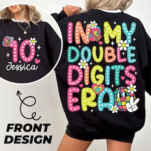 May include: Black sweatshirt with a colorful design. The front features a pink number 10 with a butterfly and the name "Jessica". The back says "IN MY DOUBLE DIGITS ERA" with a disco ball graphic and flowers.