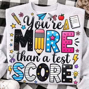 Puede incluir: Una sudadera gris claro con el texto "You're MORE than a test SCORE" en un dise&ntilde;o colorido y juguet&oacute;n. El dise&ntilde;o incluye l&aacute;pices, estrellas, tijeras y una manzana, perfecto para profesores o estudiantes.