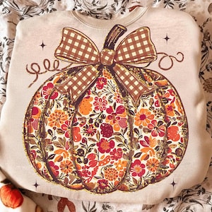 May include: Cream-colored sweatshirt featuring a large pumpkin design. The pumpkin is filled with a floral pattern in shades of red, orange, and yellow. A brown plaid bow sits atop the pumpkin, with a brown stem. The design includes decorative stars.