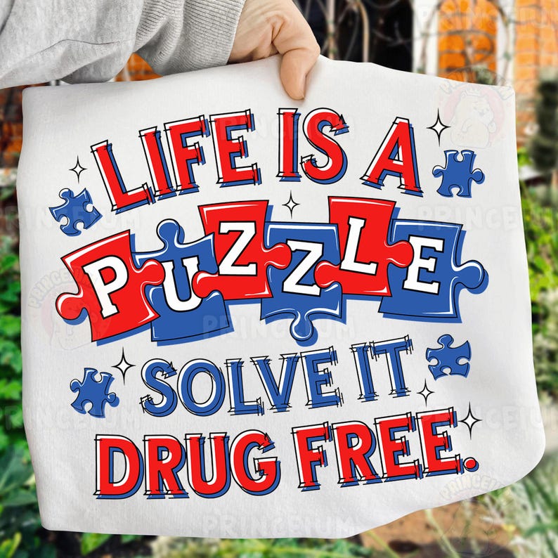 May include: White fabric with the text "LIFE IS A PUZZLE SOLVE IT DRUG FREE" in red and blue. The word "PUZZLE" is designed as puzzle pieces. The fabric is being held up by a person.