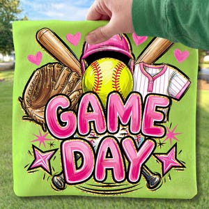 May include: A lime green square cushion featuring a softball design. The image includes a yellow softball, pink helmet, baseball bat, glove, and the words "GAME DAY" in pink. Pink hearts and stars complete the design.
