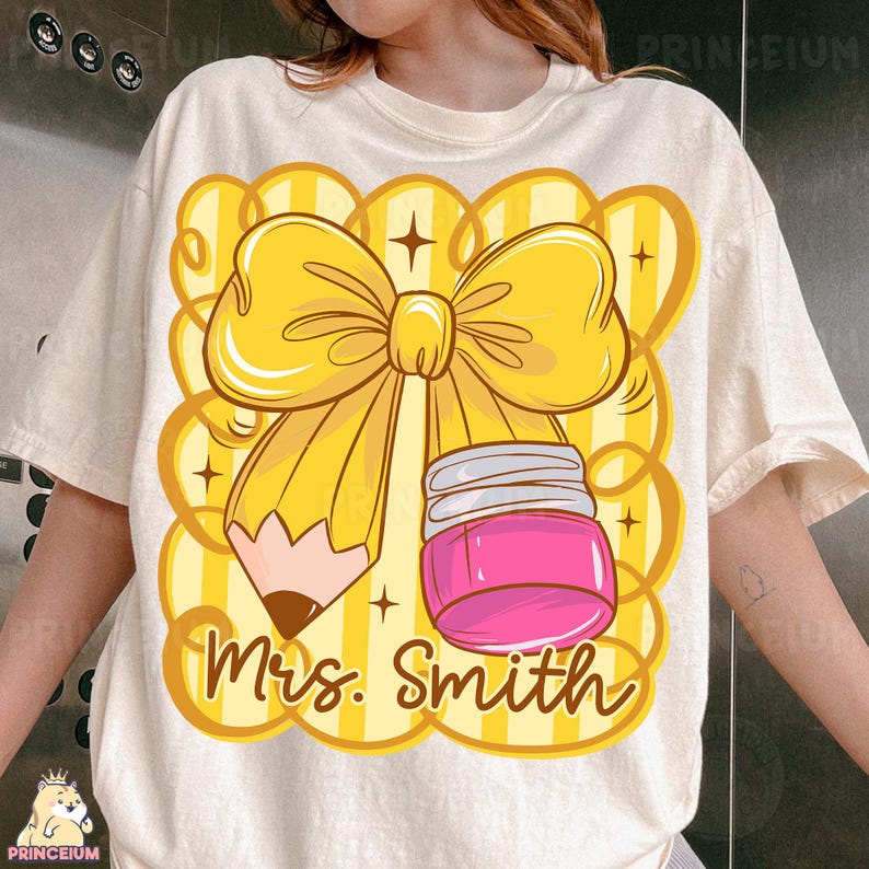 May include: Cream-colored t-shirt featuring a large graphic of a yellow bow, a pencil, and a pink jar. The pencil has a brown tip, and the jar has a silver lid. The words "Mrs. Smith" are written in a cursive font below the graphic.