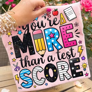 Puede incluir: Camiseta rosa claro con el texto "You're MORE than a test SCORE" en un dise&ntilde;o colorido y juguet&oacute;n. El dise&ntilde;o incluye un l&aacute;piz, estrellas y otros gr&aacute;ficos escolares.