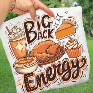 May include: White pillow with a Thanksgiving-themed graphic. The design includes a turkey, pie, cupcake, and a coffee drink. Text reads "Big Back Energy" in brown and orange lettering. The pillow is held up by a person.