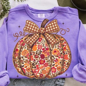 May include: A lilac Comfort Colors sweatshirt featuring a large pumpkin design. The pumpkin is filled with a floral pattern in shades of orange, red, and brown. A brown and white checkered bow sits atop the pumpkin, with the word "gee" in cursive.