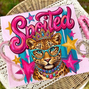 Op de afbeelding: Roze T-shirt met een luipaardafbeelding met een kroon en bril, het woord "Spoiled" in roze script en steraccenten. De luipaard is versierd met een juwelenketting. Het shirt is omgeven door sieraden en haarclips.