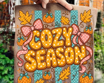May include: A brown t-shirt featuring the text COZY SEASON in orange and white checkered lettering. The design includes orange autumn leaves, small orange pumpkins with blue stems, and two large pink bows with white polka dots on a striped background.