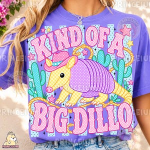 May include: A purple t-shirt with a graphic featuring a cartoon armadillo wearing a cowboy hat, surrounded by cacti and stars. The text reads "KIND OF A BIG DILLO" in pink bubble letters, with a rope design.